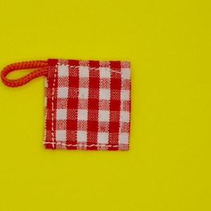 Puede incluir: Una pequeña etiqueta cuadrada de tela con un patrón de cuadros rojos y blancos, sujeta a un lazo de cordón naranja. La etiqueta está sobre un fondo amarillo brillante.