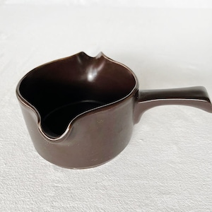 May include: A brown ceramic gravy boat with a handle. The boat has a slightly wavy rim and a smooth, glossy finish.