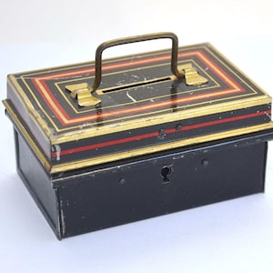 May include: A black metal money box with a gold and red striped design. It has a slot on the top and a keyhole on the front.