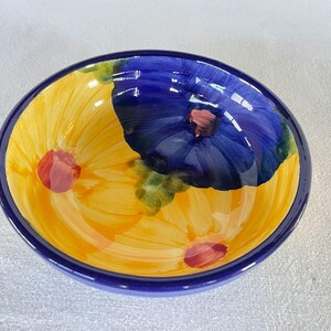 Puede incluir: Un cuenco de cerámica pintado a mano con un vibrante diseño floral. El cuenco presenta un interior amarillo con grandes flores estilizadas en amarillo, azul y rojo. El borde es de un azul oscuro, creando un contraste llamativo.
