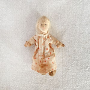 May include: A small, handmade doll with blonde hair and a pink and white dress. The doll is lying on a white fabric background.