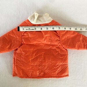 May include: An orange quilted jacket with a cream-colored ribbed collar. The jacket is approximately 14 inches long, as measured with a ruler. The jacket has a diamond pattern and is likely for a baby or toddler.