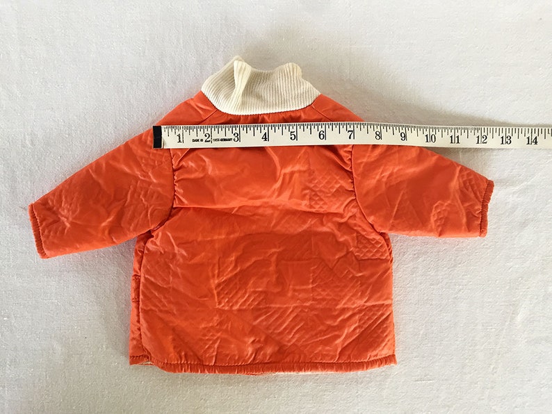 May include: An orange quilted jacket with a cream-colored ribbed collar. The jacket is approximately 14 inches long, as measured with a ruler. The jacket has a diamond pattern and is likely for a baby or toddler.