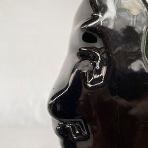 K&ouml;nnte beinhalten: Eine schwarze Keramikskulptur eines Gesichts mit einer gro&szlig;en Nase und einem geschlossenen Mund. Die Skulptur ist gl&auml;nzend und hat eine glatte Oberfl&auml;che.