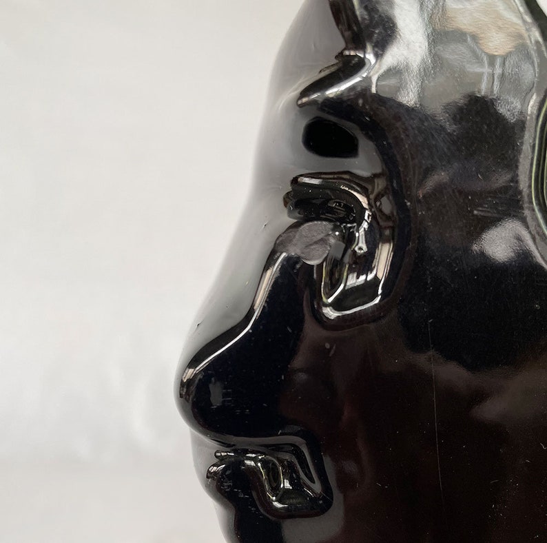 K&ouml;nnte beinhalten: Eine schwarze Keramikskulptur eines Gesichts mit einer gro&szlig;en Nase und einem geschlossenen Mund. Die Skulptur ist gl&auml;nzend und hat eine glatte Oberfl&auml;che.