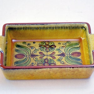 May include: A yellow ceramic baking dish with a floral design in green, brown, and red. The dish has two handles on the sides.
