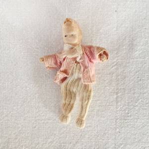May include: A small, handmade cloth doll wearing a pink and white striped sweater and pants. The doll has a simple face with blue eyes and a stitched smile.
