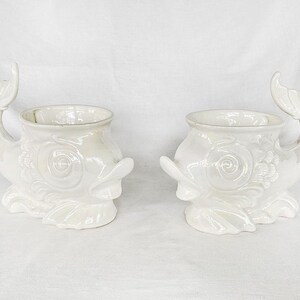 Peut inclure: Deux pots de fleurs blancs en forme de dauphin. Chaque pot a une finition nacrée, un visage de poisson et une poignée en forme de queue. Les pots ont une ouverture ronde pour les plantes et conviennent à l'intérieur ou à l'extérieur.