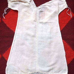 May include: A white cotton baby gown with lace trim around the neckline, sleeves, and hem. The gown has a simple, classic design.