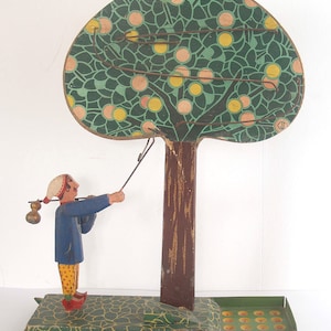 May include: A vintage wooden toy game featuring a painted figure of a person with a basket, attempting to catch apples on a wire track attached to a green and brown tree. The game has a green base with a metal tray for the apples.