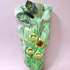 May include: A ceramic wall vase with a green and white glaze. The vase is decorated with a painted butterfly and yellow flowers.