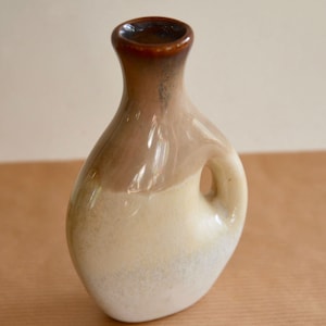 May include: A small, ceramic vase with a brown, beige, and white glaze. The vase has a rounded body and a narrow neck. It has a small handle on the side.