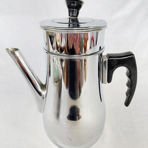 Könnte beinhalten: Ein glänzender, chromfarbener Kaffee-Perkolator mit schwarzem Griff und Deckelknopf. Der Perkolator hat einen Ausguss und einen abgerundeten Körper, der zum Brühen von Kaffee bestimmt ist. Der Griff hat ein kleeblattartiges Design.
