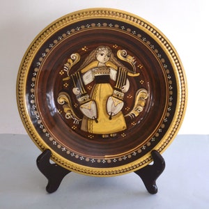 Könnte beinhalten: Ein brauner Keramikteller mit goldfarbenem Rand und einem bemalten Design einer Frau in einem gelben Kleid, die zwei weiße Gegenstände hält. Der Teller steht auf einem schwarzen Ständer.