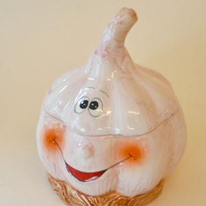 Pode incluir: Um recipiente de cerâmica branca em forma de alho, com uma base marrom e um rosto pintado. O recipiente tem uma tampa e é decorado com um rosto sorridente com olhos grandes e lábios vermelhos.