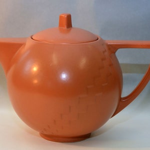 Könnte beinhalten: Eine terracottafarbene Teekanne mit rundem Korpus und quadratischem Deckel. Die Teekanne hat ein strukturiertes Design an der Seite und einen Griff, der sich nach oben und über die Oberseite krümmt.