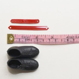 May include: Two pairs of black leather doll shoes with a white sole, two red plastic straps, and a pink measuring tape with the words "Fiber-Glass" and "China" printed on it.