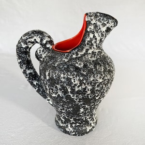 May include: A black and white ceramic pitcher with a textured surface and a red rim. The pitcher has a unique, organic shape with a wide mouth and a handle.
