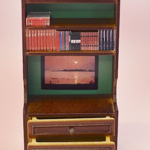 Op de afbeelding: Een miniatuur houten boekenkast met twee laden en een plank vol boeken. De plank heeft ook een afbeelding van een zonsondergang boven een watermassa.