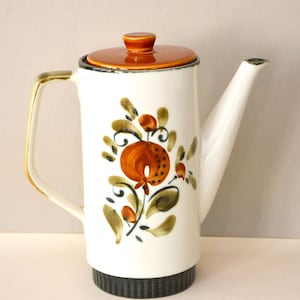 Może przedstawiać: Biały ceramiczny dzbanek do kawy z brązową pokrywką i kwiatowym wzorem w odcieniach brązu, zieleni i pomarańczy. Dzbanek ma czarną obwódkę u dołu.