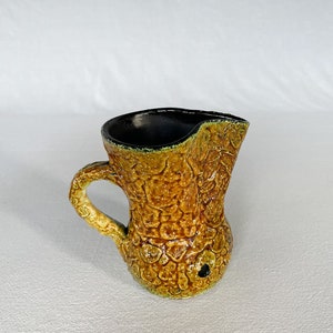May include: A yellow and green ceramic pitcher with a textured surface and a black interior. The pitcher has a handle and a spout.