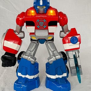 May include: A red, blue, and silver toy robot with a Transformers logo on its chest. The toy has a blue helmet, red torso, and blue legs. The robot has a weapon attached to its right arm and lights on its head.