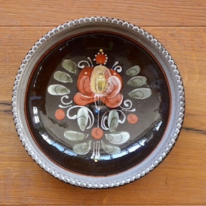 May include: A brown ceramic plate with a white rim and a hand-painted floral design. The flower is red, orange, and yellow with green leaves and a white background.