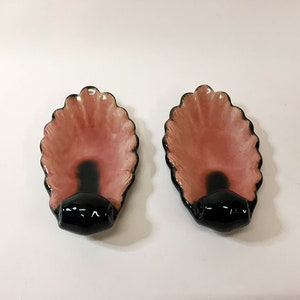 May include: Two pink and black ceramic wall hooks shaped like seashells. The hooks have a scalloped edge and a black knob on the bottom.