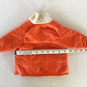 May include: Bright orange quilted jacket with a cream-colored ribbed collar. The jacket measures approximately 12 inches across the chest. The jacket is likely for a small child or pet.