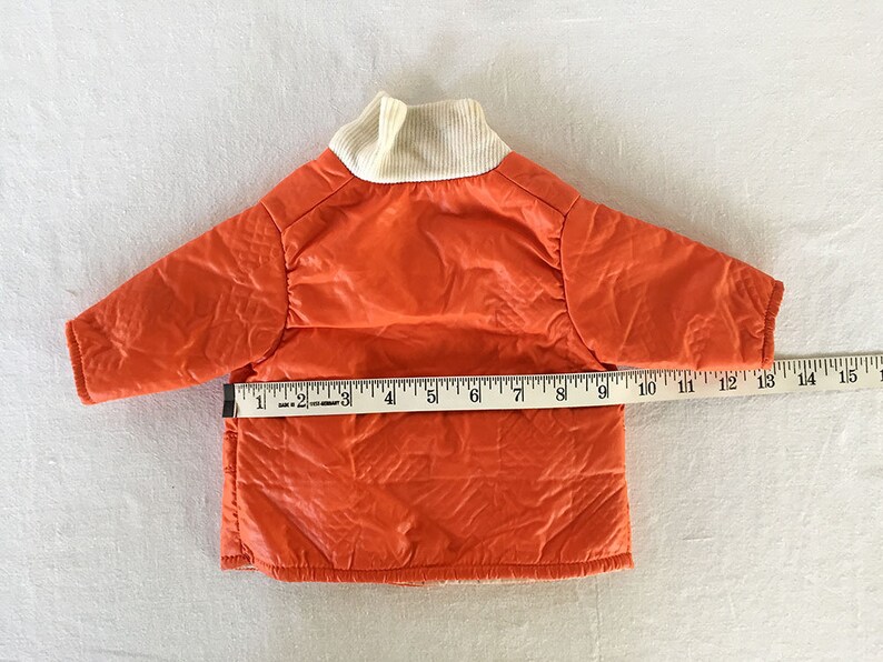 May include: Bright orange quilted jacket with a cream-colored ribbed collar. The jacket measures approximately 12 inches across the chest. The jacket is likely for a small child or pet.