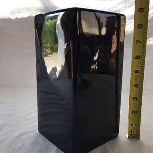 May include: A glossy black square vase with a tapered base. The vase is approximately 10 inches tall, as measured by a yellow tape measure. The surface reflects the surrounding environment.