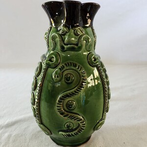 Puede incluir: Un jarrón de cerámica verde con la parte superior de color marrón oscuro y un diseño en relieve de una serpiente con una cara en la parte delantera.
