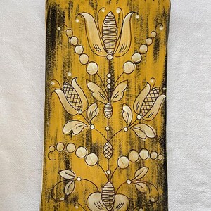 Op de afbeelding: Rechthoekige decoratieve keramische tegel met een gele achtergrond en een bloemenontwerp. Het bloemenontwerp heeft crèmekleurige bloemen, bladeren en cirkelvormige accenten, met zwarte omlijning en accenten. De tegel heeft een verweerde, getextureerde uitstraling.