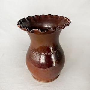 May include: A handcrafted, brown ceramic vase with a ruffled rim. The vase has a bulbous body and a decorative band of small, raised circles around the neck. The glaze has a glossy finish, and the vase is suitable for displaying flowers or as a decorative piece.