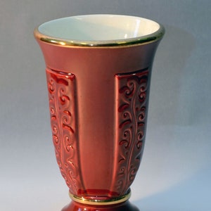 May include: A tall, red vase with a gold rim and base. The vase has a flared top and a pedestal base. Intricate, raised scrollwork adorns the sides. The interior is white.