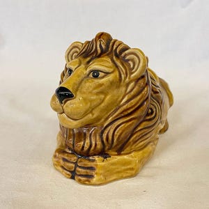 May include: A ceramic lion figurine in shades of tan and brown. The lion is depicted in a seated position with its head turned slightly to the side. The mane and facial features are detailed with darker brown accents.