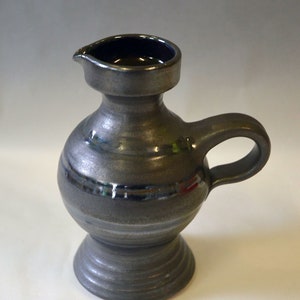 May include: A small, gray ceramic pitcher with a rounded body and a handle. The pitcher has a slightly flared lip and a base with a ring of raised lines.