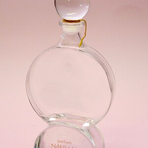 May include: A clear glass bottle with a round, clear glass stopper. The bottle has a round base and the label reads "parfum NAHEMA guerlain PARIS".