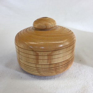 Puede incluir: Una caja de madera con tapa, la caja es de color marrón claro y tiene un acabado liso. La tapa tiene un pomo redondeado en la parte superior.