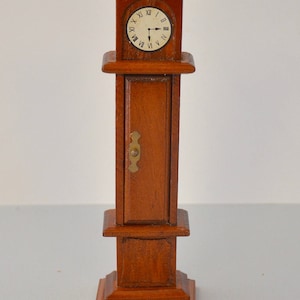May include: A miniature wooden grandfather clock with a white face and black hands. The clock is about 6 inches tall and has a small door on the front.