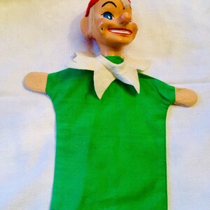 May include: A green felt hand puppet with a white collar. The puppet has a painted face with a red hat and a gold ornament on top.