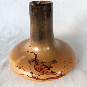 May include: A brown ceramic vase with a wide base and a narrow neck. The vase is decorated with a painted image of a bird on a branch. The artist's signature, "Anna Rio", is visible on the vase.