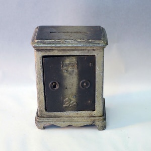 May include: A vintage cast iron piggy bank shaped like a small safe with a slot on top and a door that opens. The safe is painted silver with black accents and has a keyhole and a number "22" on the door.
