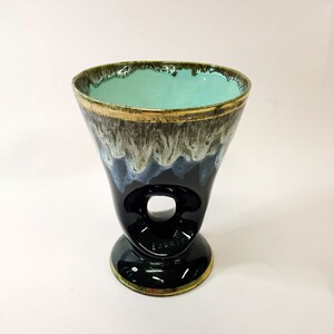 May include: A black ceramic vase with a teal glaze and gold trim. The vase has a unique shape with a hole in the side and a flared base. The vase is marked "Lourdes" on the bottom.