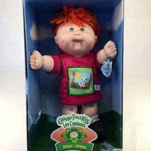 Könnte beinhalten: Eine Cabbage Patch Kids Puppe in einem rosa Shirt mit einem bunten Patch und schwarzen Schuhen. Die Puppe hat rotes Haar und grüne Augen. Die Puppe befindet sich in einer durchsichtigen Plastikbox mit grünem und blauem Hintergrund. Die Box trägt das Cabbage Patch Kids Logo und die Worte "Kid Enfant Peperini".