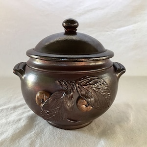 May include: A brown ceramic pot with a lid, featuring a raised design of leaves and fruit. The pot has two handles and a rounded shape.