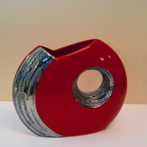 May include: A red ceramic vase with a silver design. The vase has a circular opening in the side.