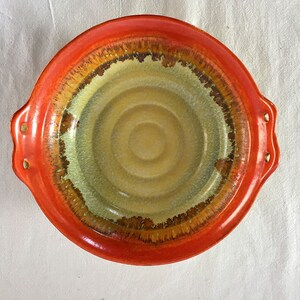 May include: A round orange ceramic bowl with two handles. The bowl has a light brown and green glaze with a concentric circle pattern.