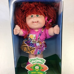 Könnte beinhalten: Eine Cabbage Patch Kids Puppe mit roten Wollhaaren, einem rosa Oberteil und einem bunten, gemusterten Rock. Die Puppe befindet sich in einer durchsichtigen Plastikbox mit blauem und grünem Hintergrund. Die Box trägt das Cabbage Patch Kids Logo und den Text "Kid Enfant Peperini".