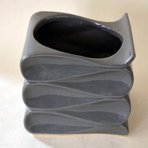 May include: Gray ceramic planter with a wavy, stacked design. The planter has a wide opening and a smooth, glossy finish.